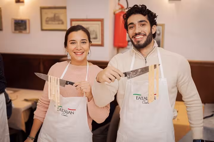 Smiling couple holding knives with fresh pasta at a cooking class in Rome's Piazza Navona, wearing aprons.