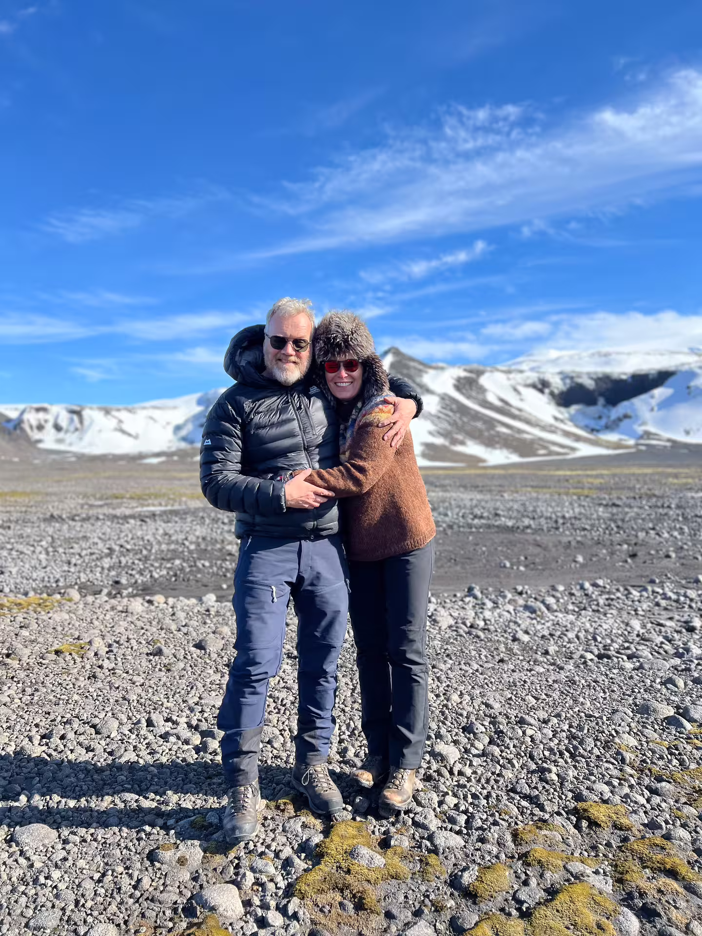 Couple embracing on rocky glacier terrain with snow-capped mountains in Iceland on a clear sunny day