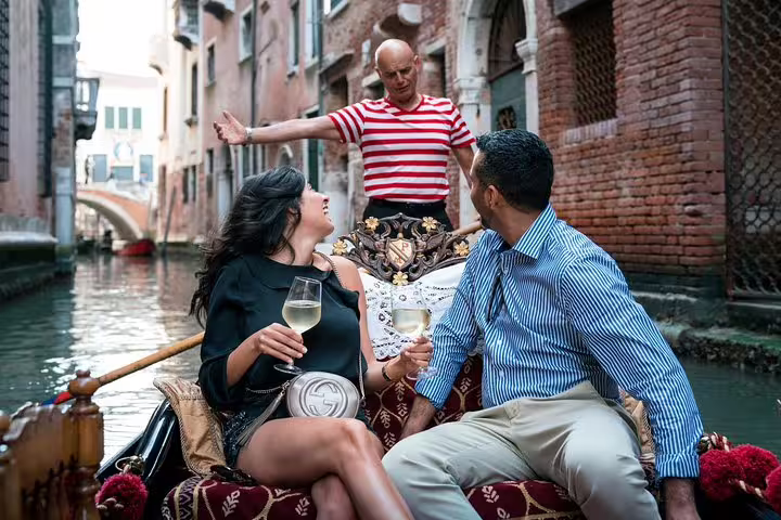 Couple enjoying a gondola ride in Venice with a gondolier, sipping wine and taking in the historic canal scenery.