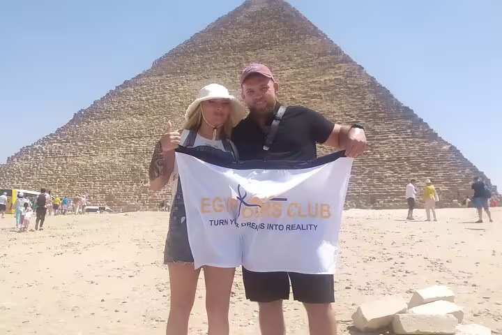 Couple holding Egypt Tours Club banner in front of the Great Pyramid of Giza on a half-day tour
