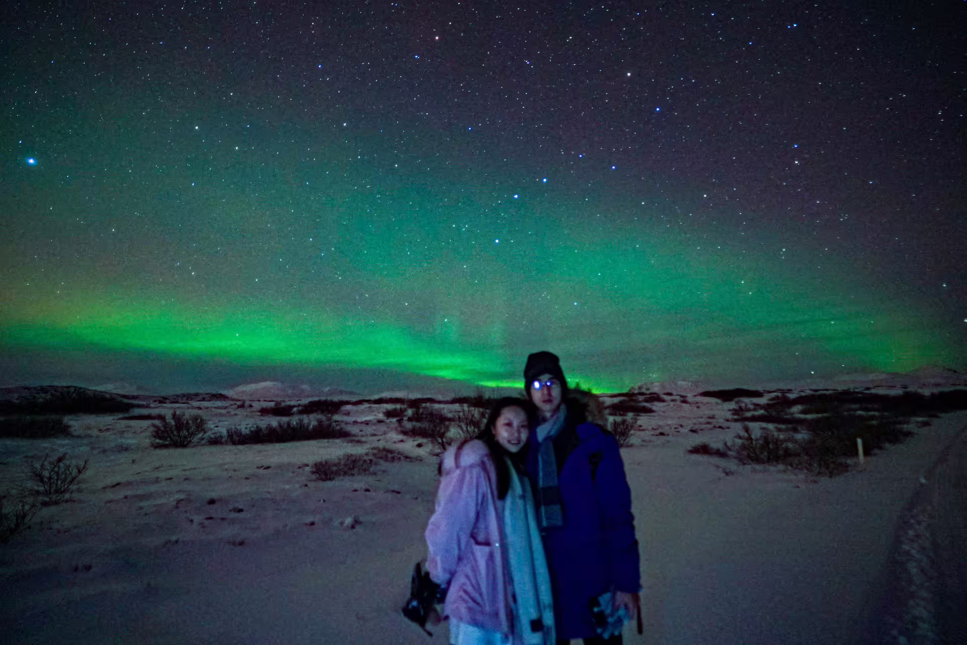 Couple posing under vibrant green northern lights aurora borealis on a snowy Iceland landscape near Reykjavik.