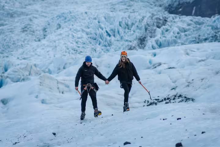 A couple holding hands walks across a glacier wearing crampons and helmets during an adventurous private glacier hike.