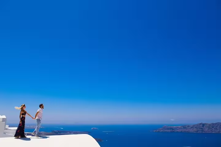 Couple holding hands on a bright Santorini rooftop with a panoramic view of the deep blue Aegean Sea.