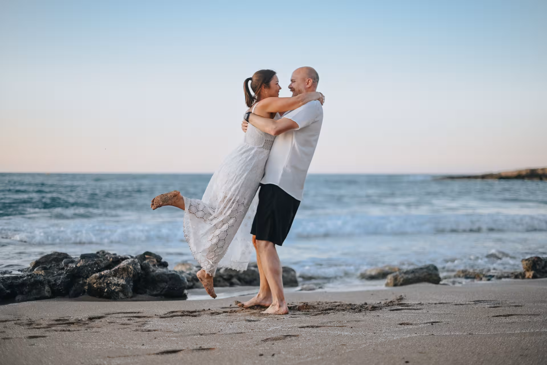 Joyful couple playfully hugging on a beach with waves and rocks at the Port of Hersonissos.