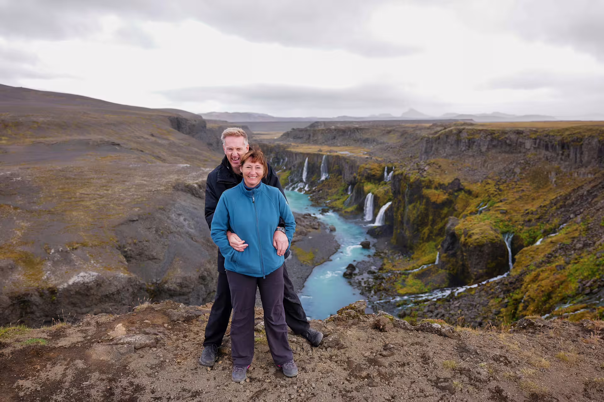 Smiling couple posing at Iceland hidden highlands canyon overlook with turquoise river and cascading waterfalls below