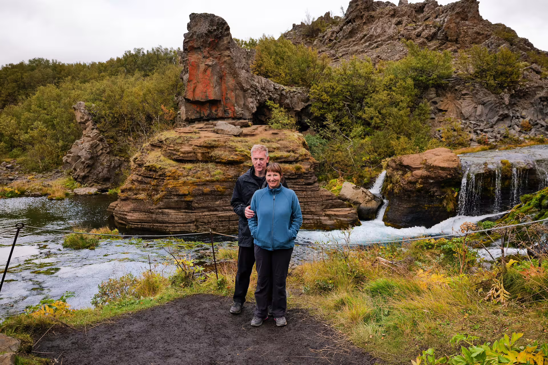 Couple standing by a stunning Iceland highland waterfall with layered red volcanic rocks on The Hidden Highlands tour