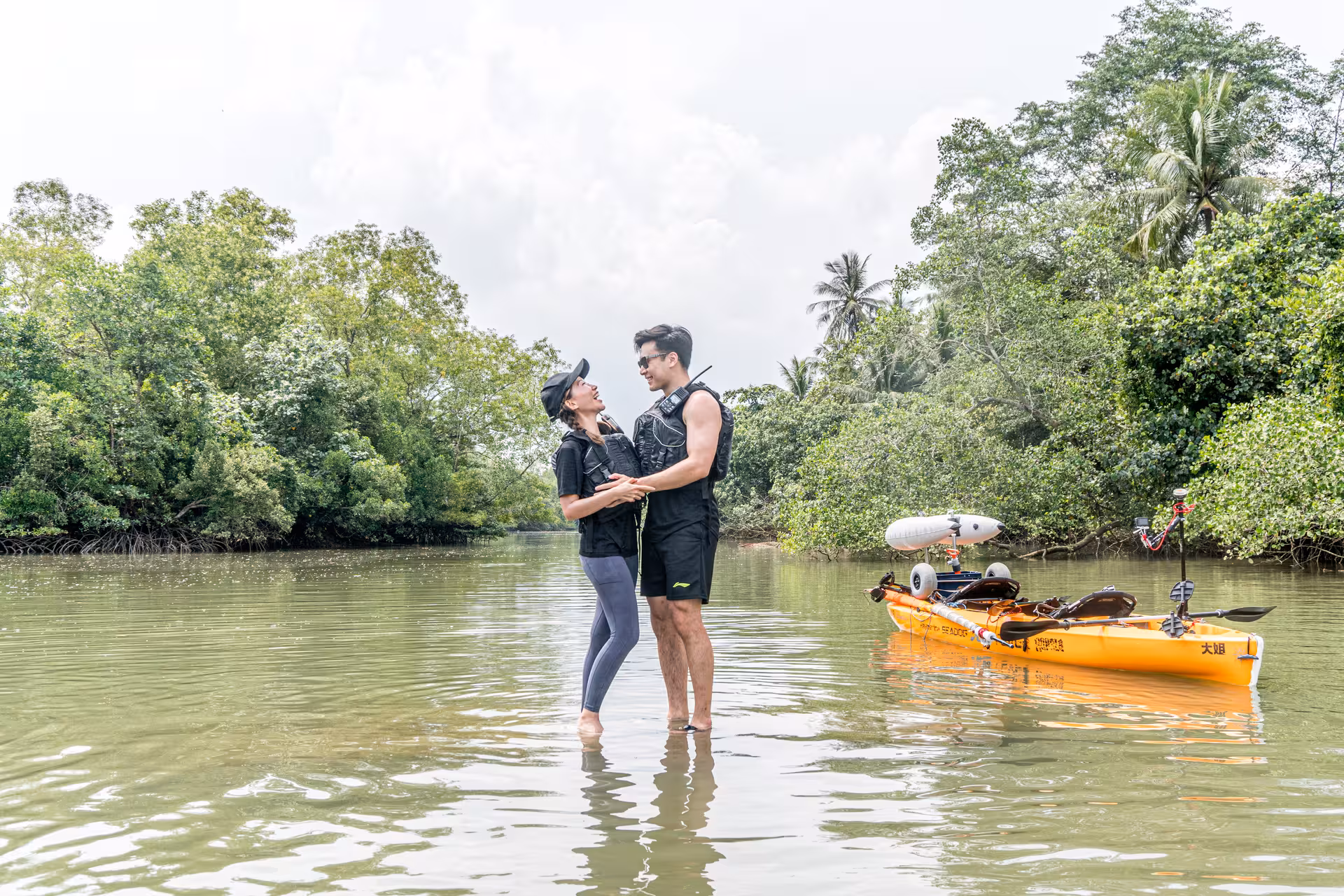 Couple standing in shallow water, smiling near a kayak, amidst lush greenery during their kayak sailing tour.