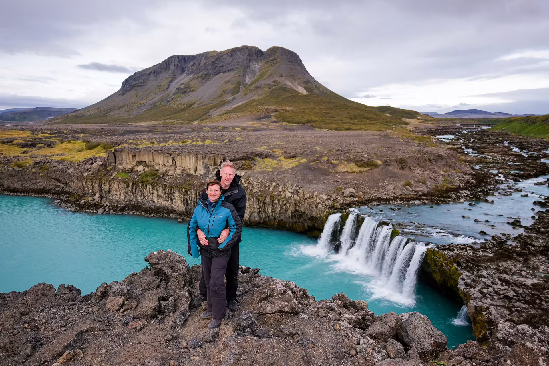 Happy couple poses on rocky Iceland highland cliff overlooking a turquoise waterfall and towering volcanic mountain on a scenic tour.