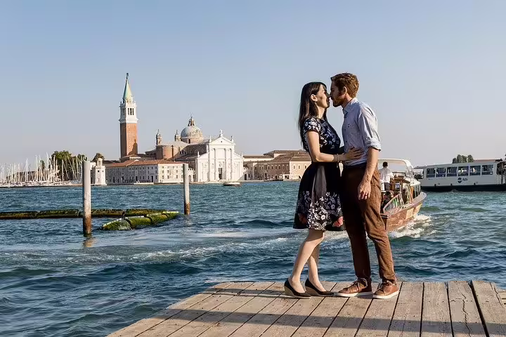 Couple sharing a kiss on a pier with iconic Venetian landmarks and a boat on the Grand Canal in the background.