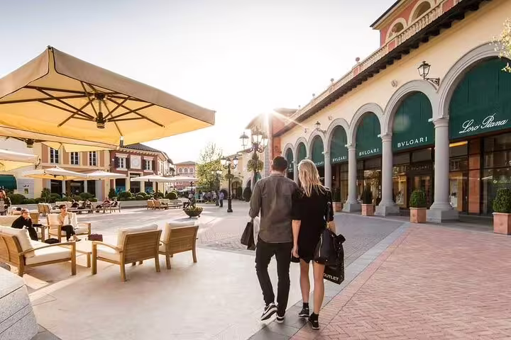 Couple strolling through a luxury shopping plaza in Milan, featuring designer stores and outdoor seating under large umbrellas.