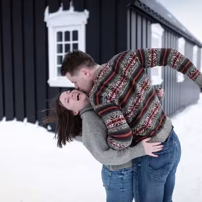 A joyful couple in Nordic sweaters sharing a romantic moment outside a black Icelandic building in snowy Snaefellsnes
