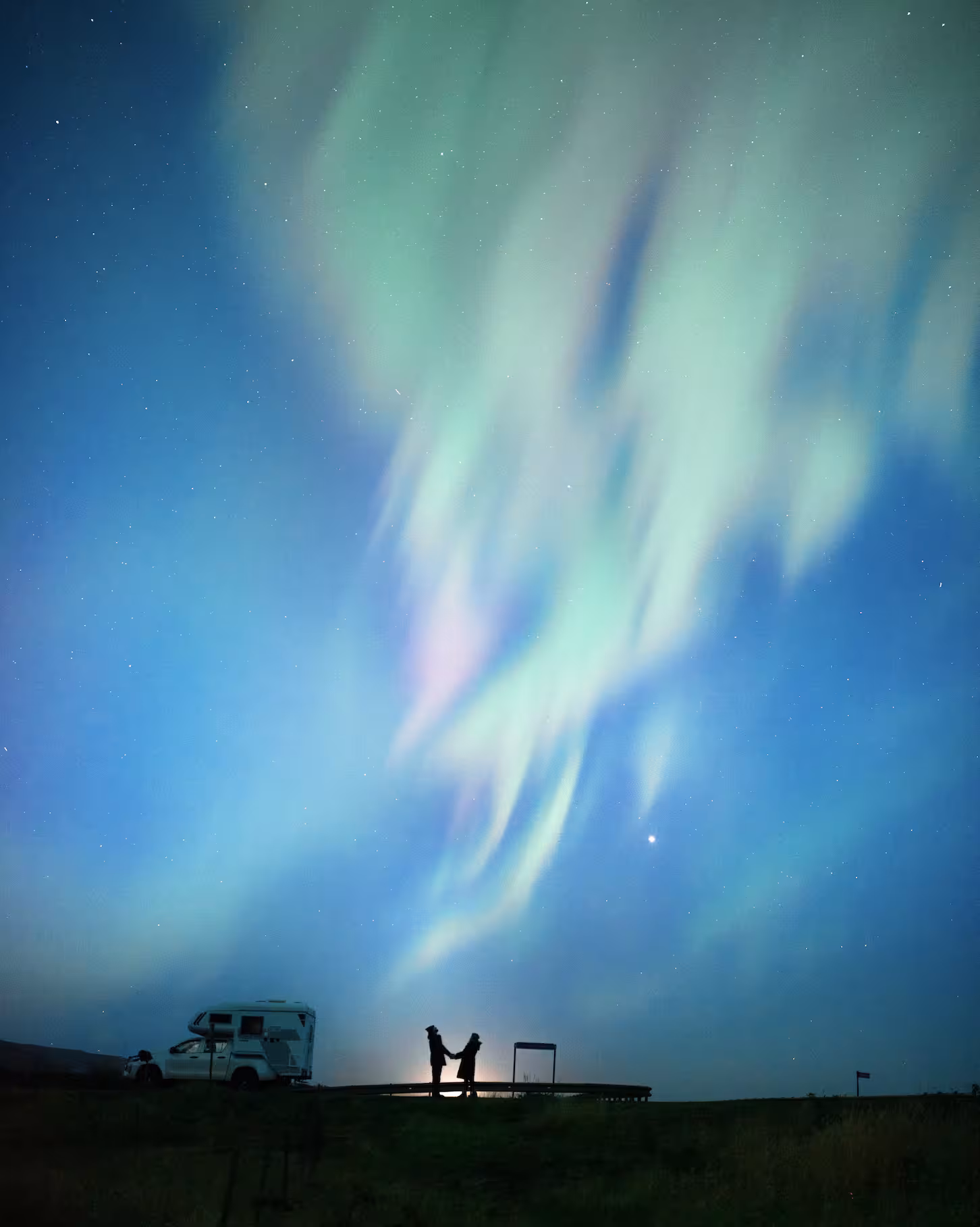 Romantic couple silhouetted beneath vibrant green and pink northern lights aurora borealis in Iceland night sky.