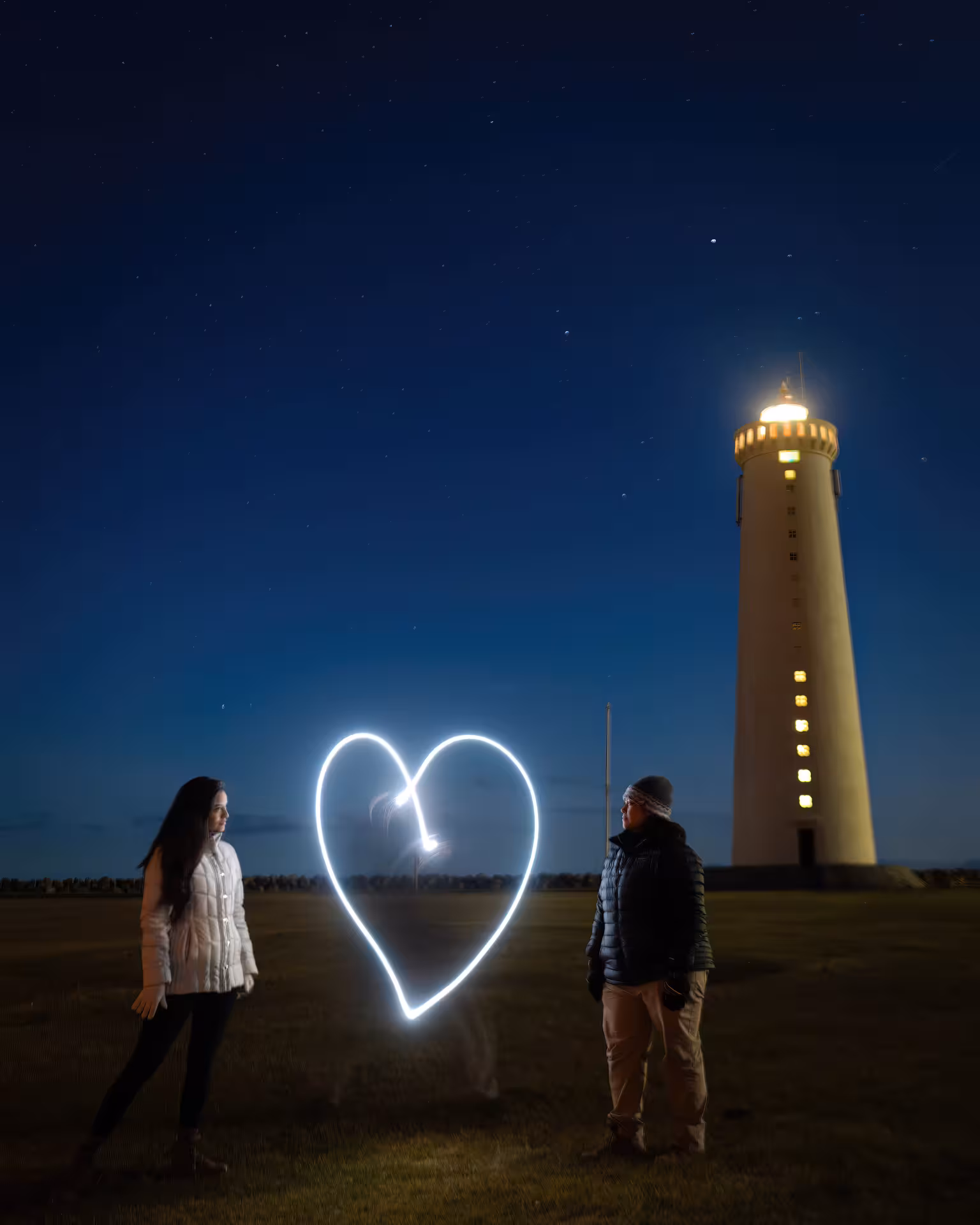 Couple standing by a glowing light-painted heart near an Iceland lighthouse under a stunning starry night sky