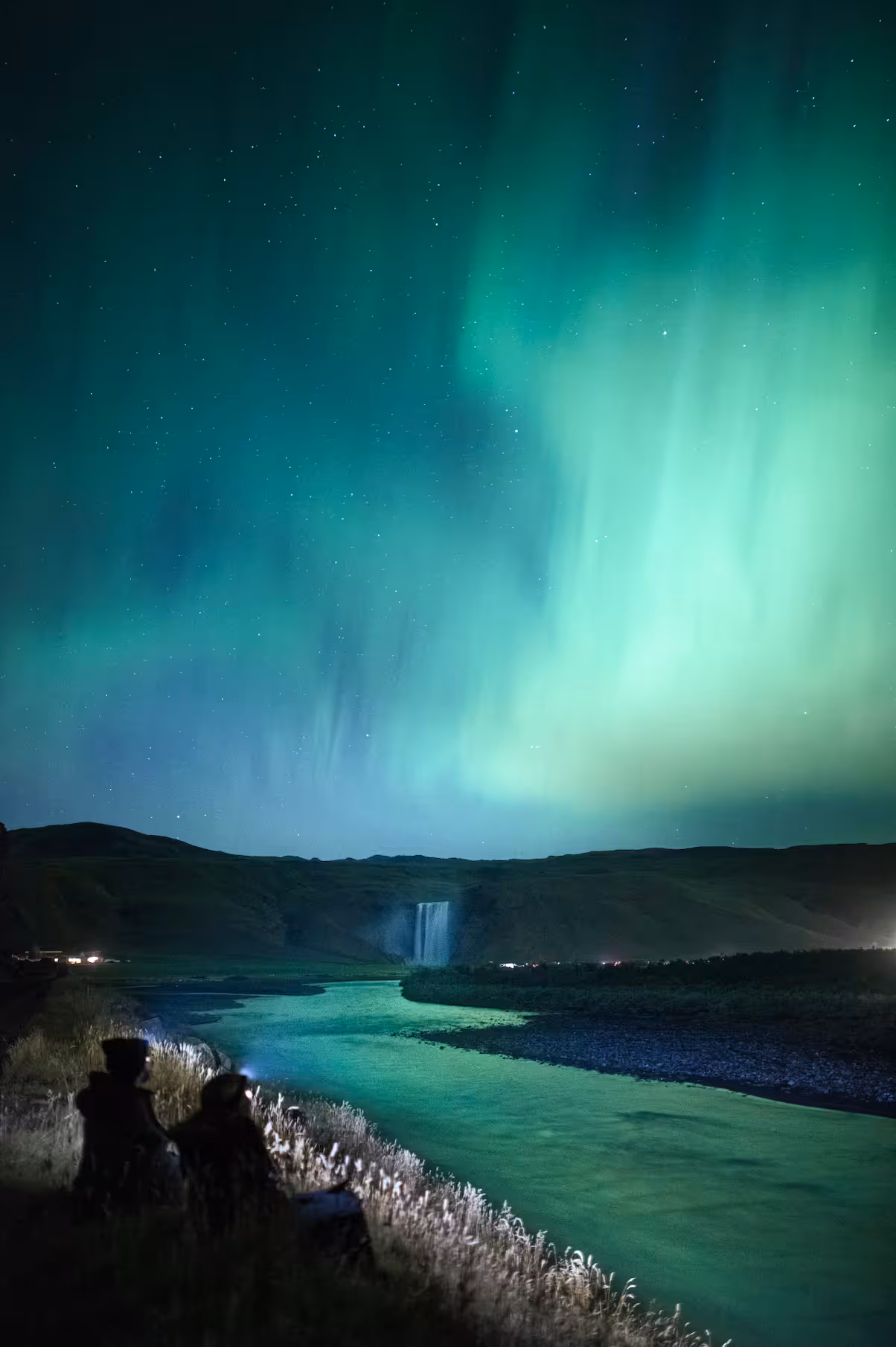 Silhouetted couple watching vibrant green aurora borealis dance over an Icelandic river and waterfall at night.