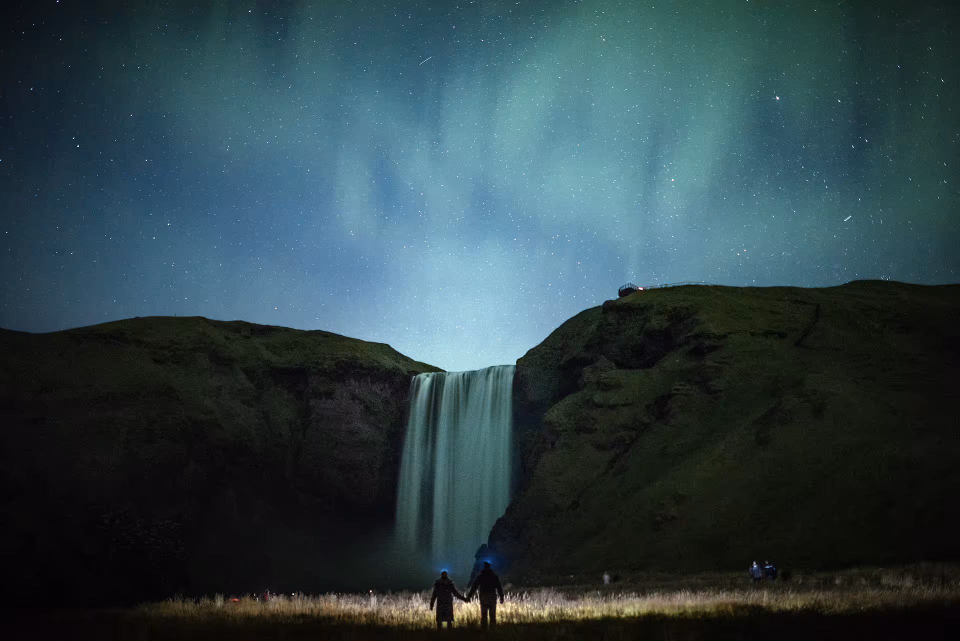 Couple holding hands beneath Iceland's Skogafoss waterfall with glowing northern lights and stars filling the night sky.