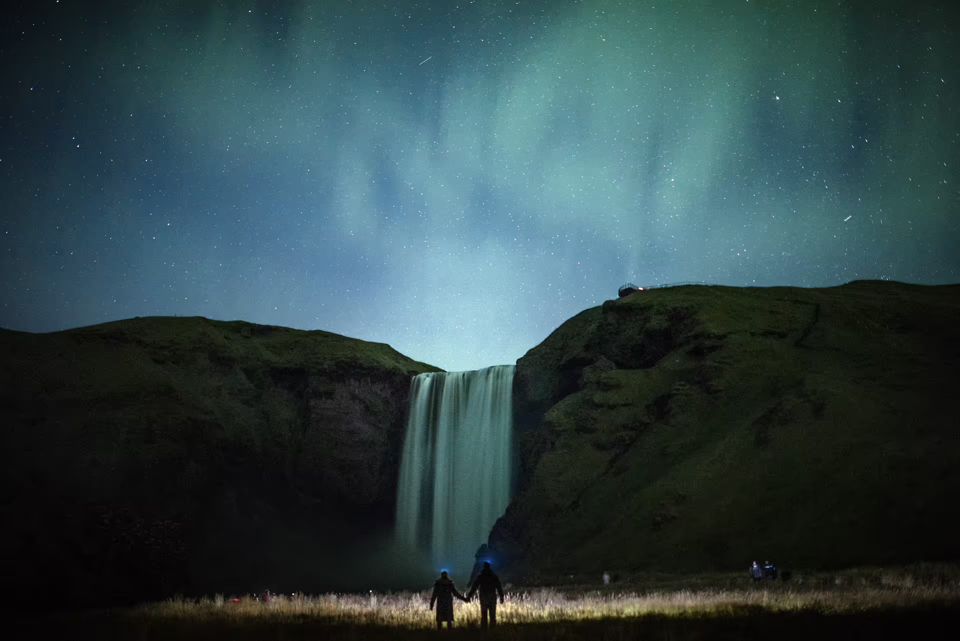 Couple holding hands beneath glowing northern lights and Skogafoss waterfall on a private Iceland aurora tour