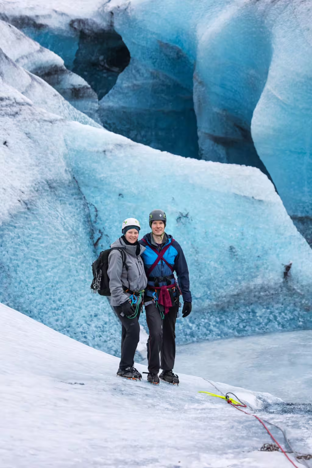 Smiling couple in helmets and harnesses stand before blue glacial ice walls on a private glacier tour