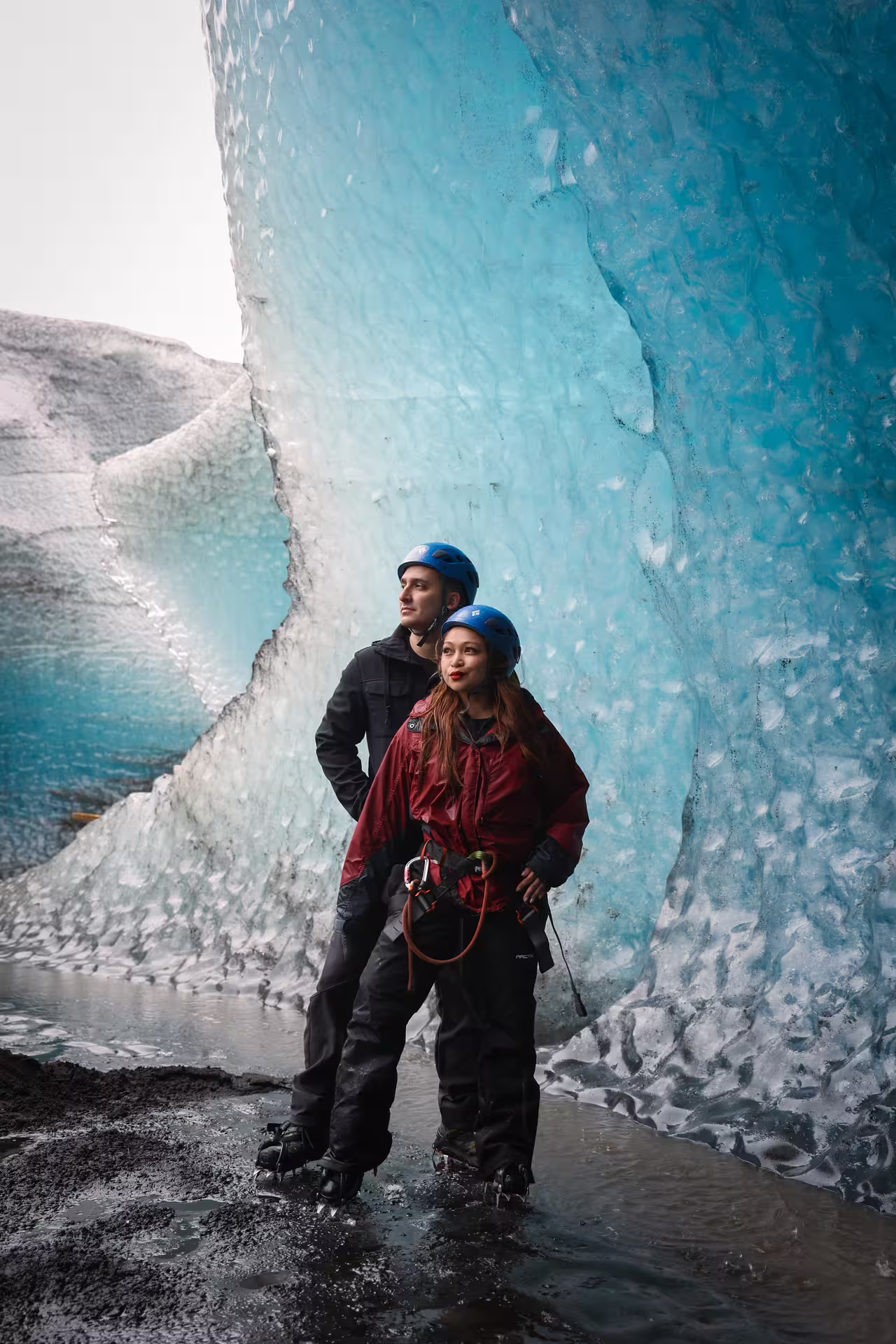 Adventurous couple in helmets and harnesses posing beside towering blue glacier walls on private tour