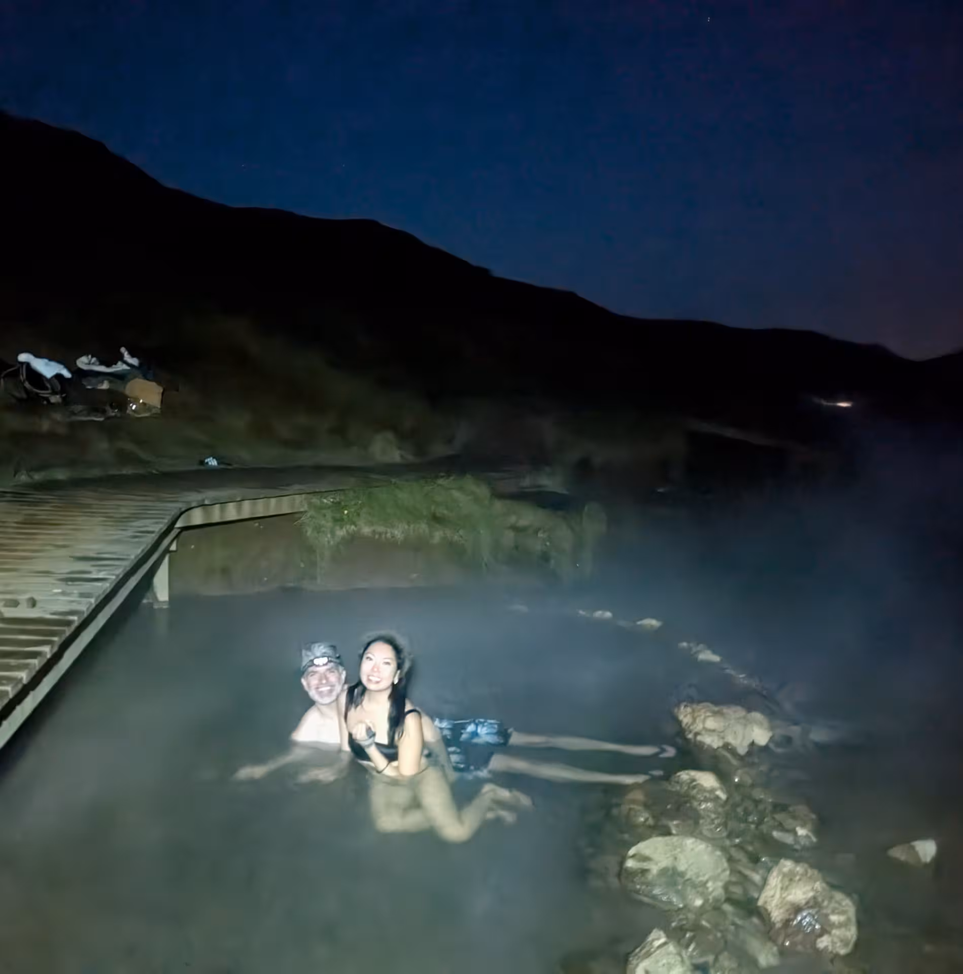 Couple soaking in steaming Reykjadalur hot river pool at night with misty hills backdrop