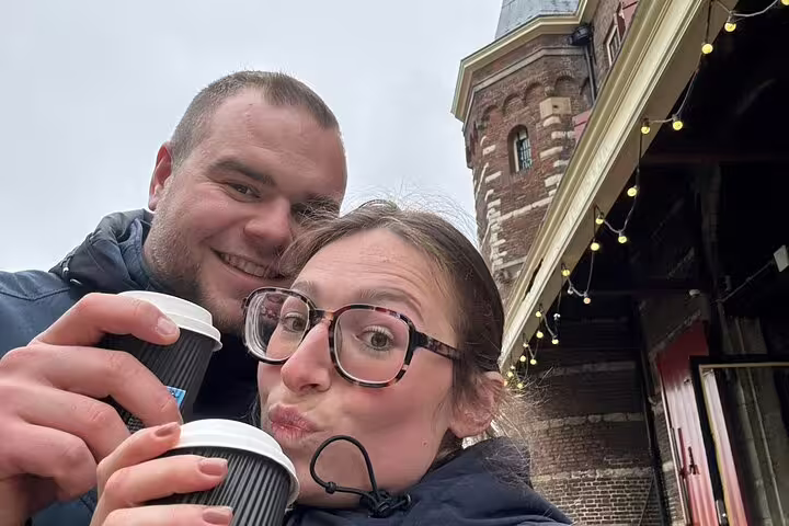 Couple taking a selfie with coffee cups in front of a historic Amsterdam brick building with string lights