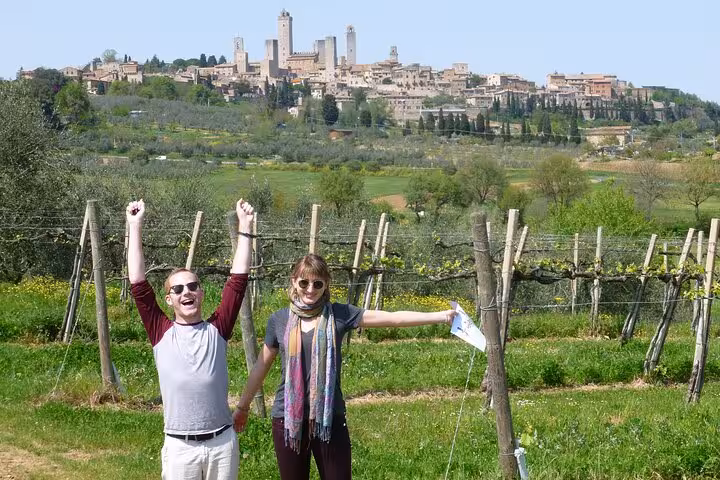 Couple celebrating in front of San Gimignano's iconic skyline, surrounded by vineyards on a sunny day in Tuscany.