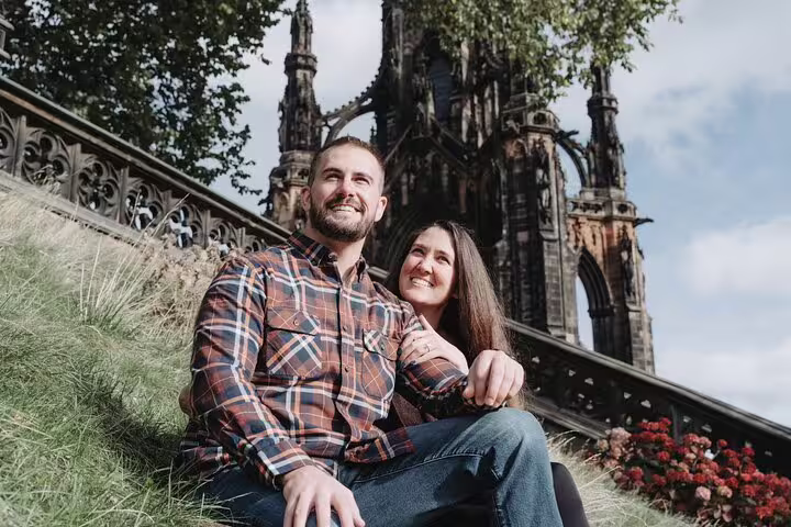 Smiling couple seated on grass with the iconic Scott Monument in Edinburgh in the background.