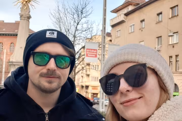 Couple wearing sunglasses and winter hats on a Budapest street with historic buildings