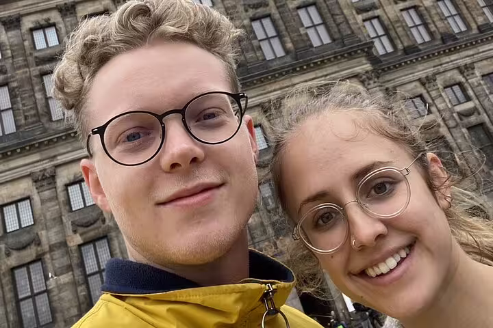Two smiling tourists wearing glasses take a selfie in front of a historic Amsterdam building