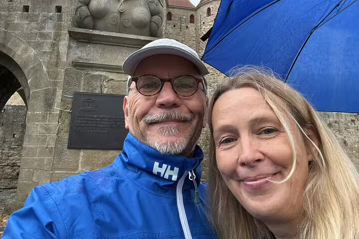 Couple smiling in front of Carcassonne medieval city walls on rainy day with blue umbrella