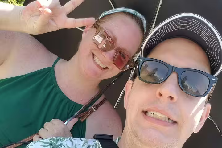 Couple taking a selfie outdoors smiling, woman flashing peace sign wearing green top and sunglasses