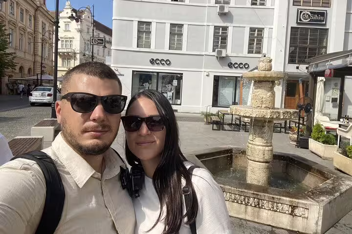 Couple taking selfie by historic stone fountain in Sibiu Old Town square with ECCO store in background