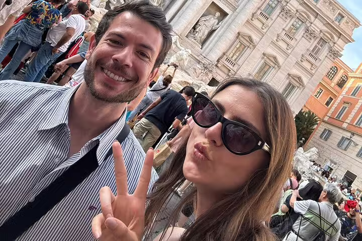 Couple taking a selfie with peace sign at Trevi Fountain Rome, crowded tourist landmark in background