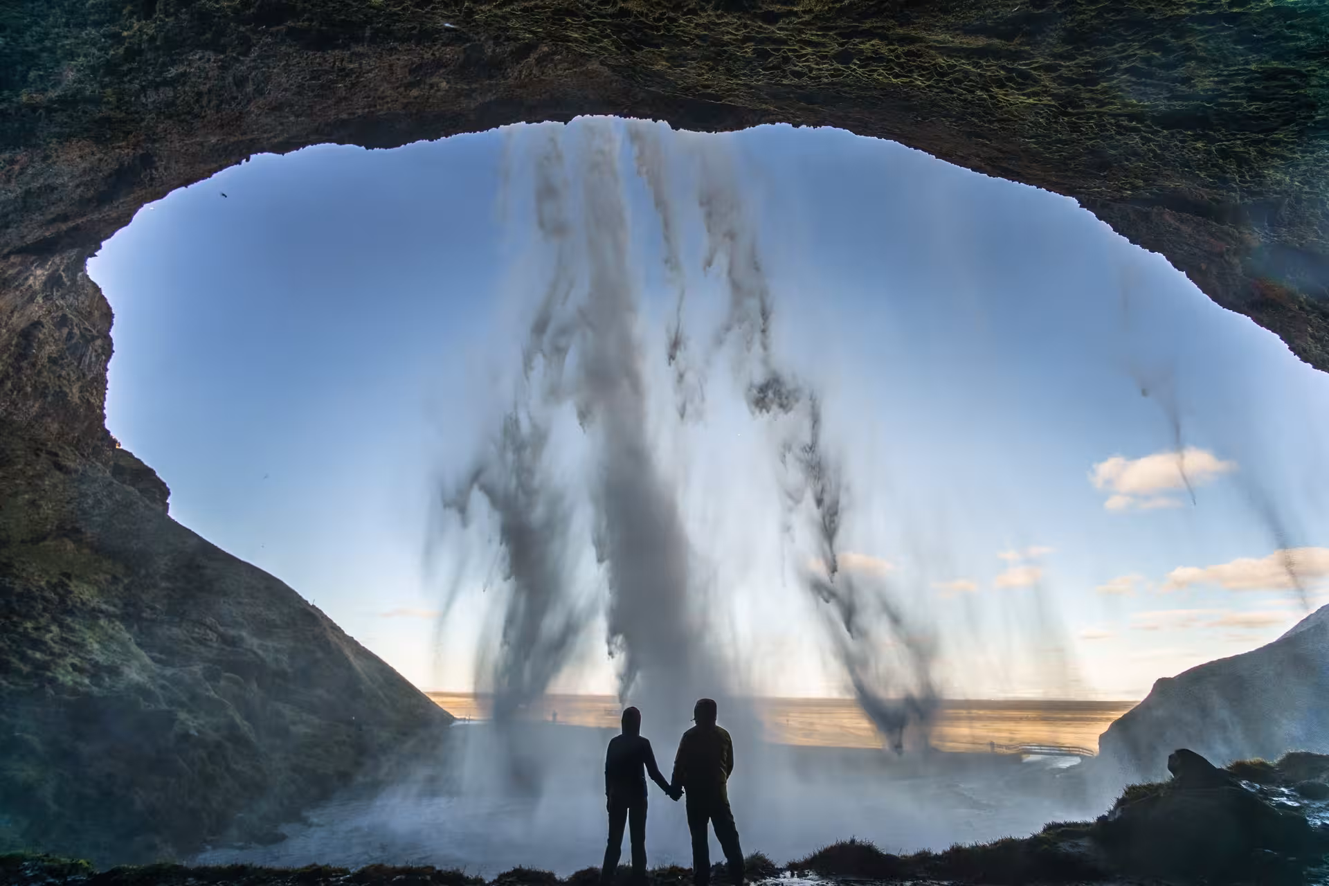 Couple holding hands behind Seljalandsfoss waterfall at sunset on Iceland 6-Day Stopover Tour.