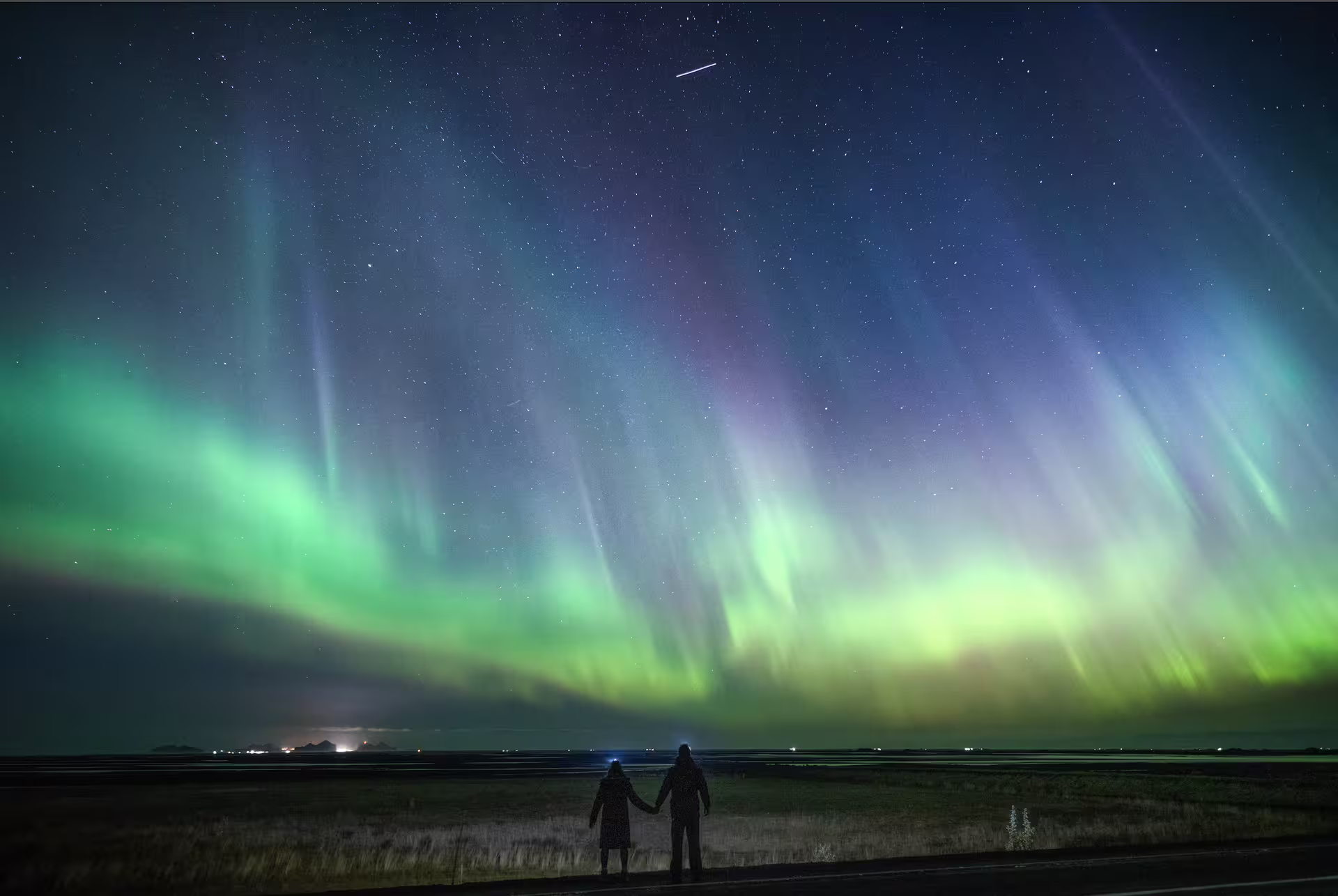 A couple holds hands silhouetted beneath dramatic green and purple northern lights on a private Iceland aurora photography tour