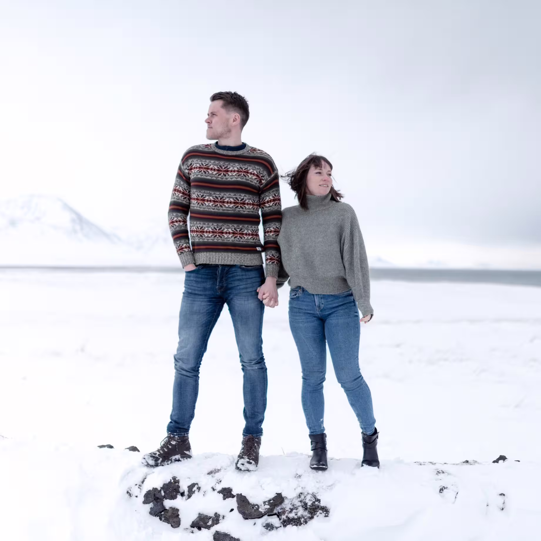 A couple in cozy knitwear standing hand in hand on a snowy Snaefellsnes landscape with misty mountains and frozen sea