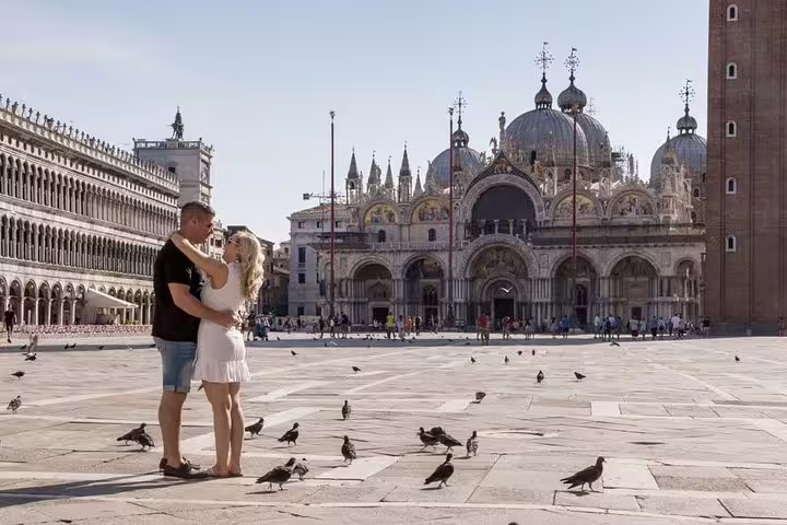 Couple embracing in St. Mark's Square, Venice, with iconic Basilica and pigeons in the background.
