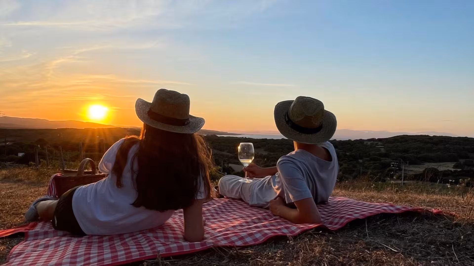 Couple enjoying a sunset picnic with wine on a checkered blanket overlooking a Gallura estate landscape.