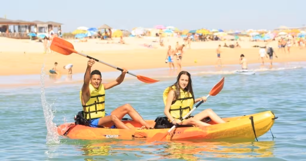 Smiling couple paddling a tandem kayak in calm turquoise water near a lively summer beach full of umbrellas and sunbathers