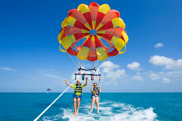 Couple enjoying tandem parasailing over turquoise ocean, flying under red and yellow parachute on boat tow