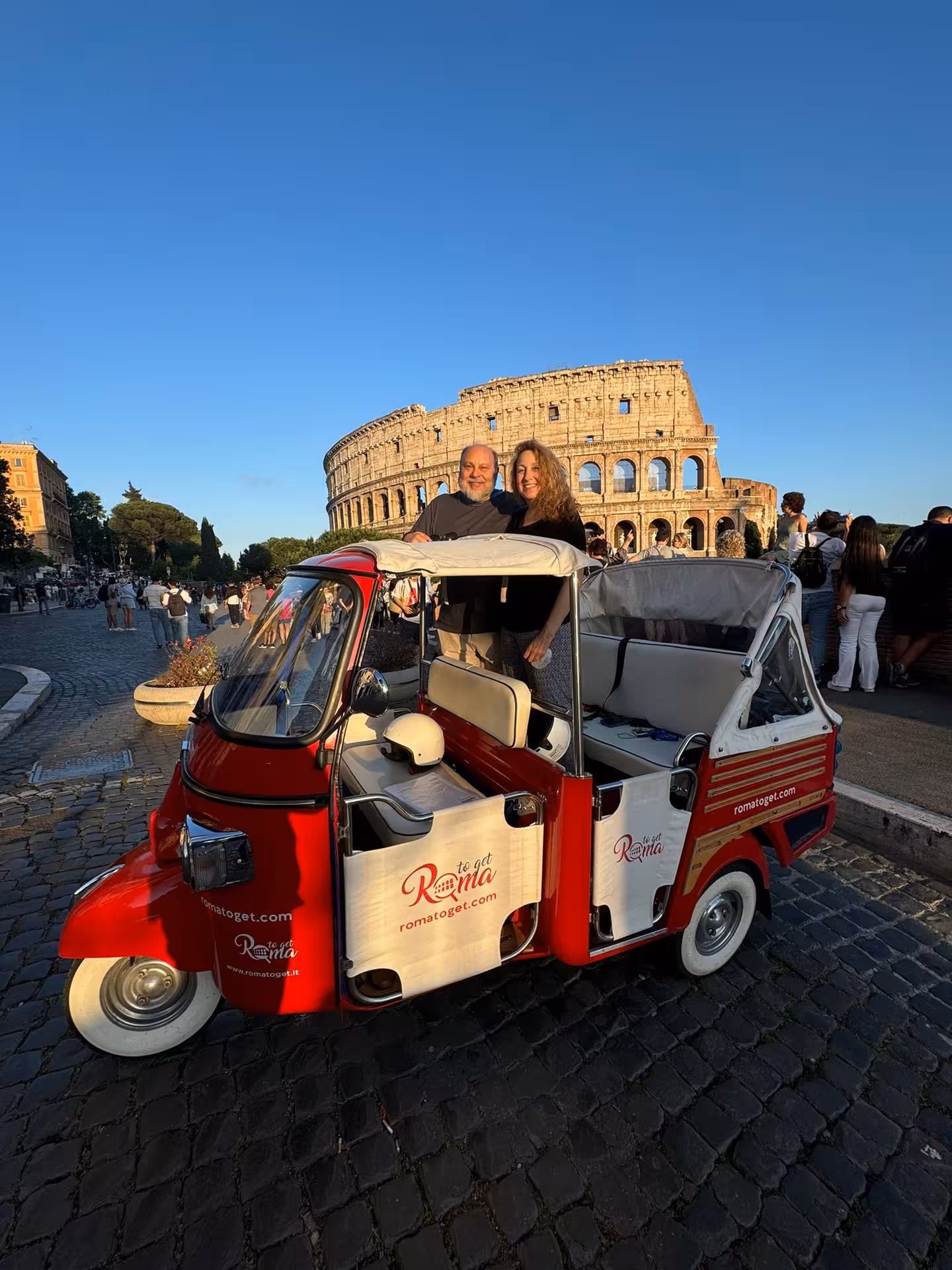 Couple enjoys a tuk-tuk tour with the Colosseum as a backdrop on a sunny day, perfect for Rome sightseeing.