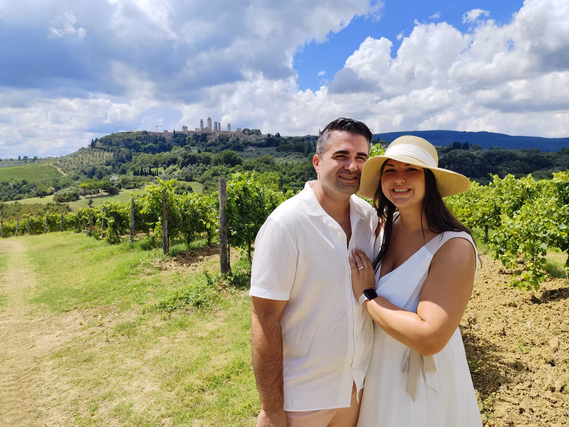 Smiling couple in a Tuscan vineyard with rolling hills and distant medieval towers under a partly cloudy sky.