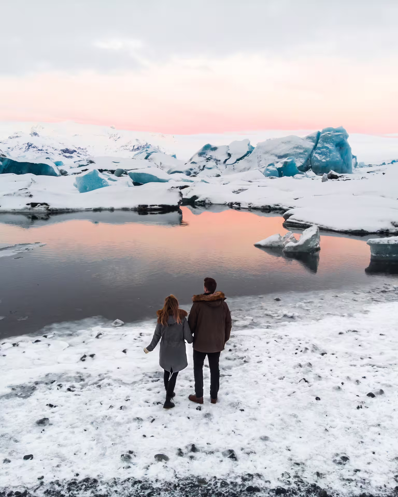 Couple holding hands at Jokulsarlon glacier lagoon with blue icebergs and pink sunset sky in Iceland