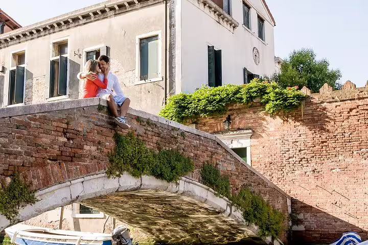 Couple embracing on a charming brick bridge in Venice, surrounded by historic buildings and lush greenery.