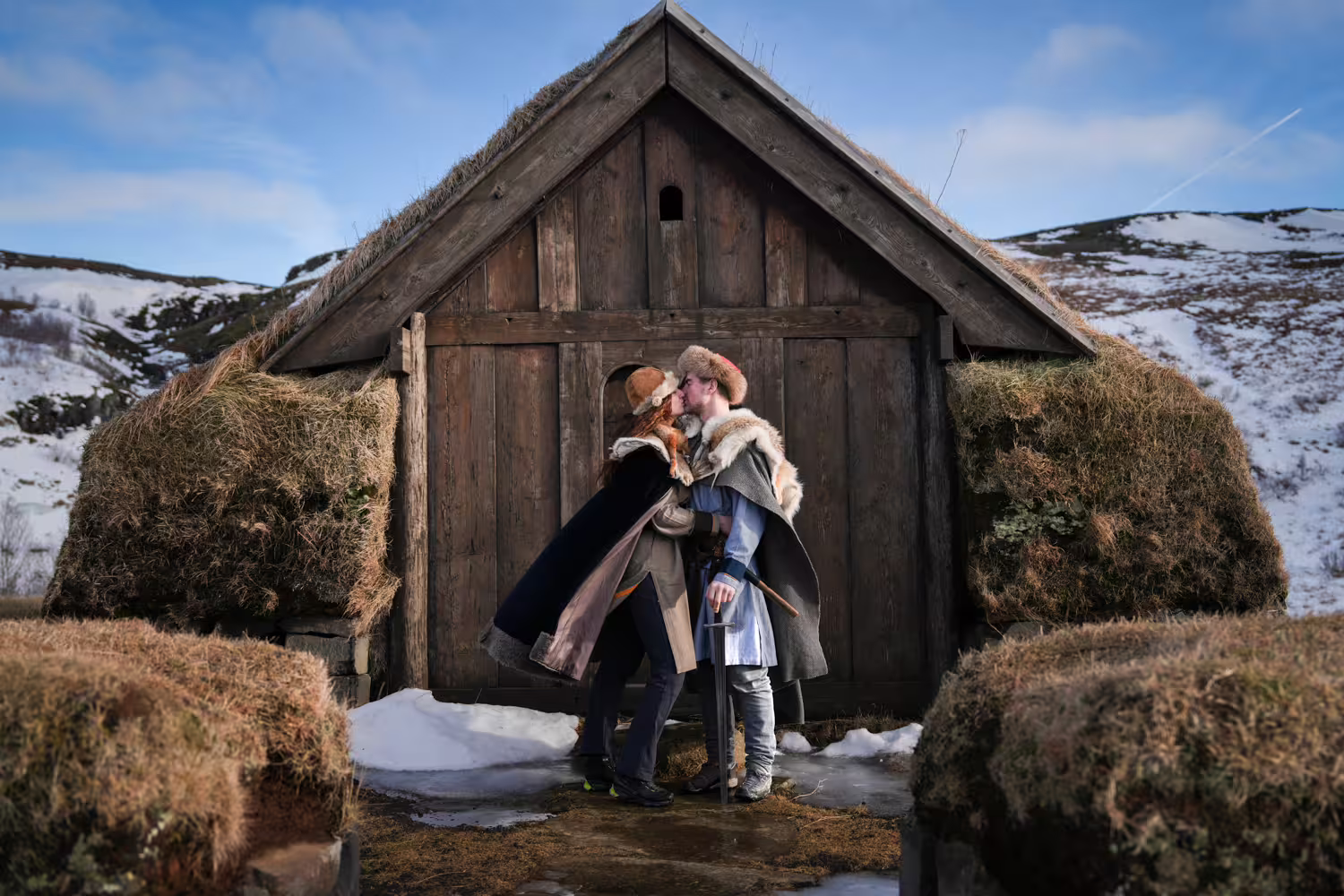 Couple in authentic Viking costumes sharing a romantic moment outside a traditional Iceland turf house in winter