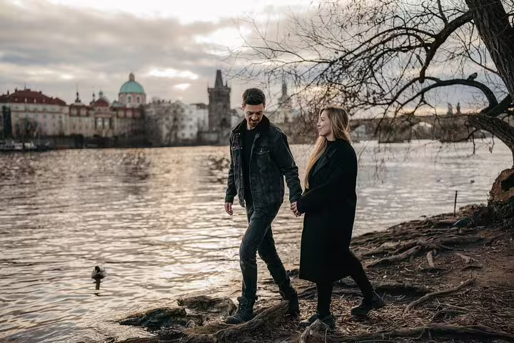 Couple walking hand in hand along the Vltava River with Prague's scenic skyline in the background during a photoshoot.