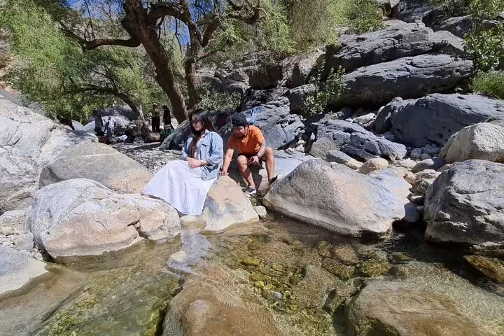 Couple enjoying serene rocky stream setting on Wadi Dham full-day tour, surrounded by lush greenery and clear waters.