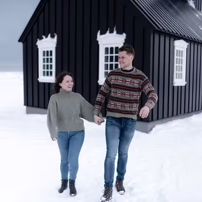 A happy couple holding hands and walking through snow past a charming black Icelandic house on the Snaefellsnes Peninsula