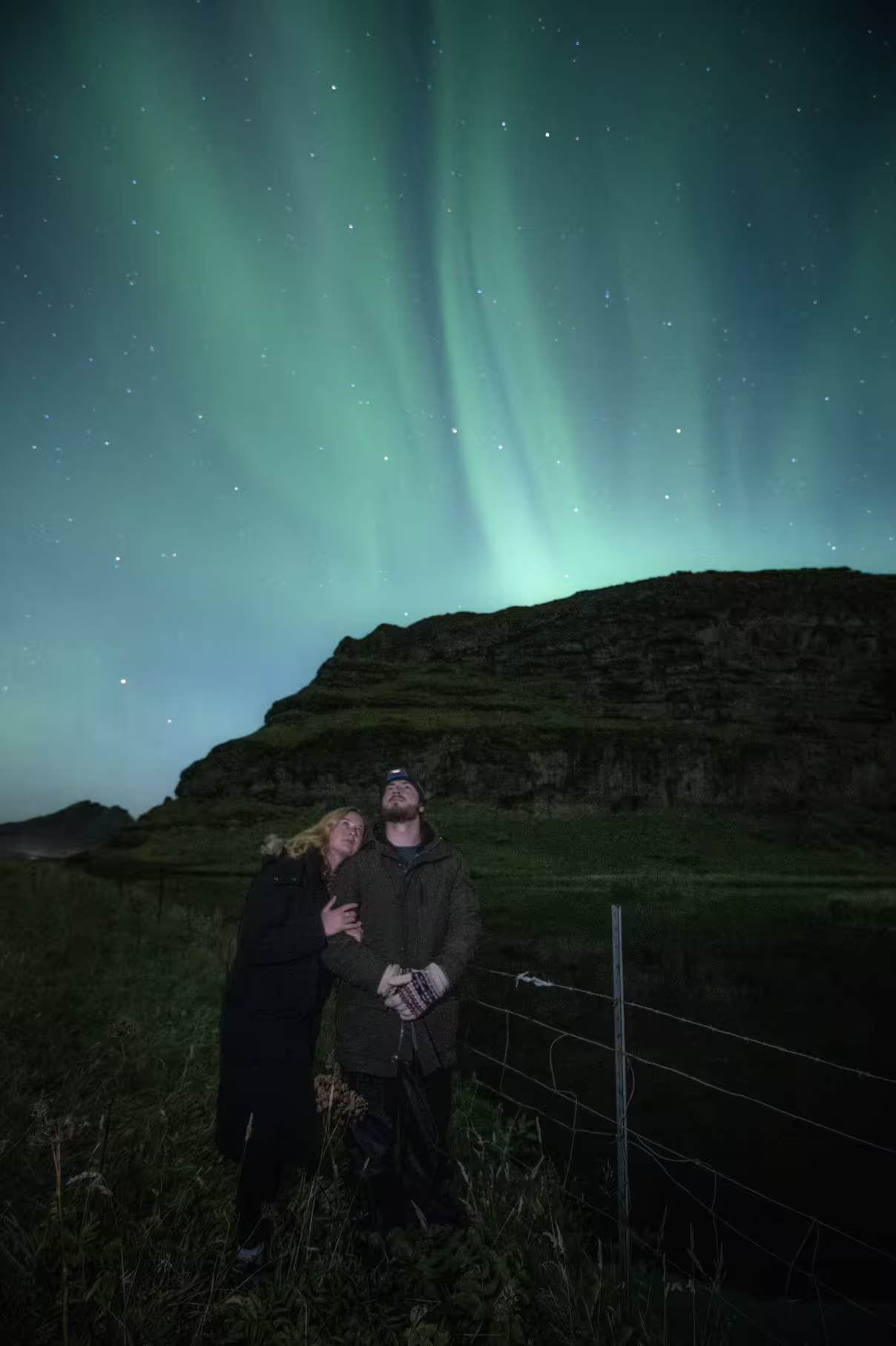 Couple embraces gazing at brilliant green northern lights over Iceland hills on a private aurora photography tour