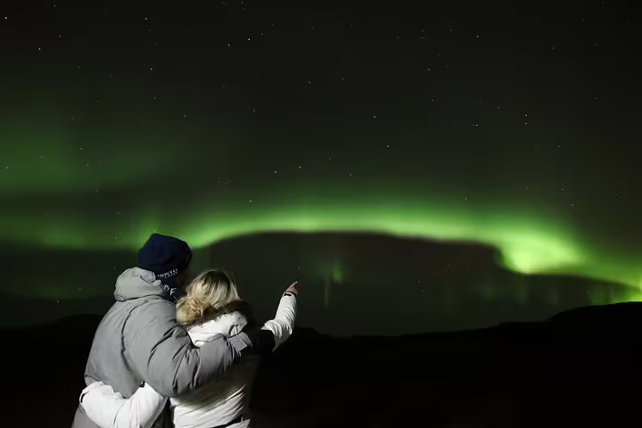 Couple embracing while pointing at vibrant green northern lights dancing across Iceland's night sky