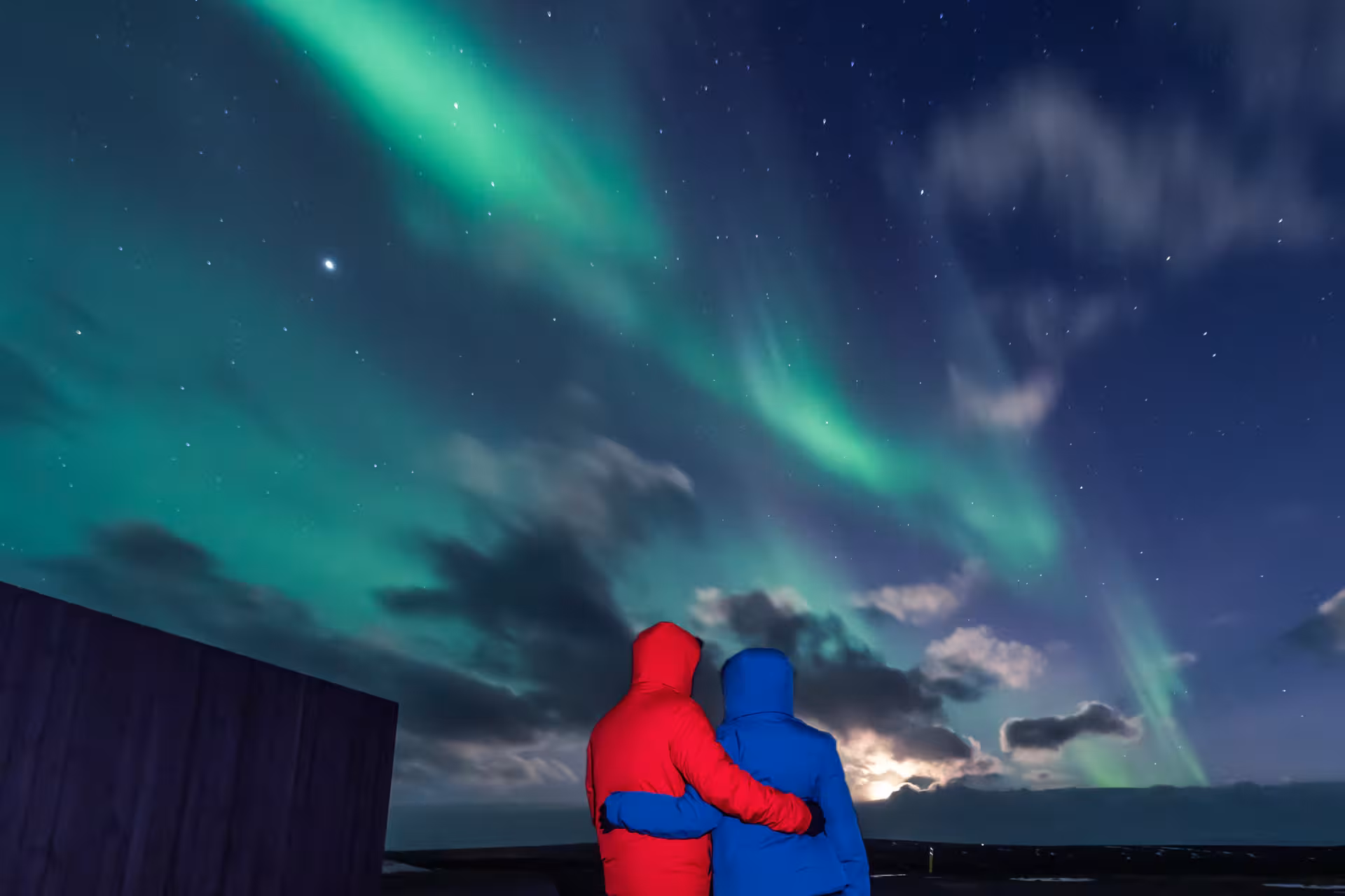 Couple in red and blue jackets embracing while watching northern lights aurora borealis over Iceland night sky